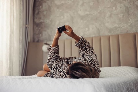 Woman in a leopard print robe lying on the bed, holding her smartphone above her. The cozy bedroom