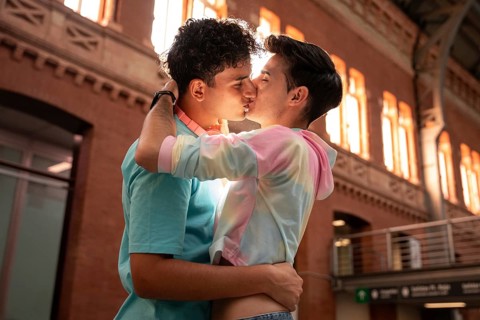 A gay couple shares an intimate kiss while wrapped in each other's arms in a warm sunlit urban
