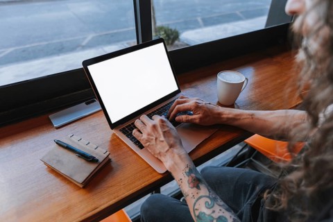 A man with tattooed arms types on a laptop at a wooden table in a cafe, creating a cozy workspace.