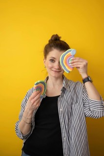 A woman joyfully holds two colorful clay rainbows against a vibrant yellow background. This