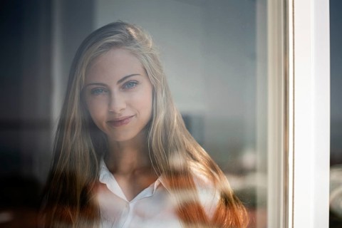 A woman with long hair gazes through a window. The glass reflects gentle light, creating a serene