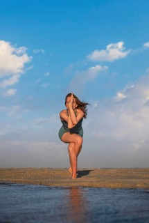 Woman stands on one leg with the other wrapped, arms crossed, practicing yoga on the beach at