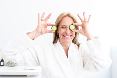 A middle-aged woman in a plush white robe holds cucumber slices over her eyes, smiling joyfully