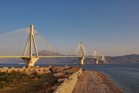 A vast bridge across the sea with beach facilities and ruins in the foreground against a mountain