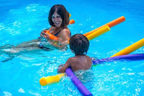 Happy mother smiling, holding a pool noodle and looking at her young son, who is also playing with
