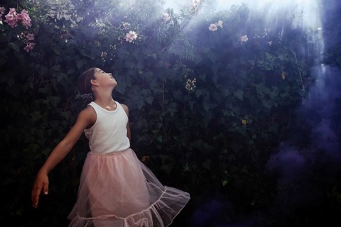 A teenage girl in a white blouse and pink skirt gazes enchanted at a lush garden, with sunlight