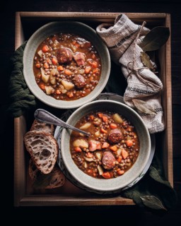 A rustic lentil stew is served in two bowls, filled with sliced sausage, carrots, and potatoes. A