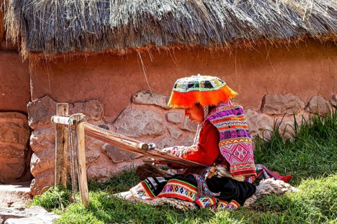 Person in traditional, colorful clothing weaving in front of a thatched hut in a meadow, elderly