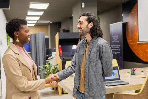 Two colleagues shake hands in a modern office. The meeting involves smiling and collaboration,