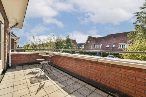 A sunny balcony with two chairs and a table overlooks a quaint neighborhood with brick houses. The