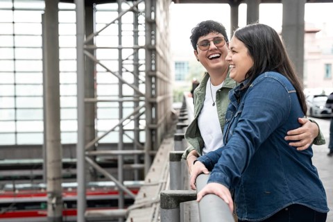 A happy lesbian couple shares a joyful moment, leaning on a railing in a train station. Both women