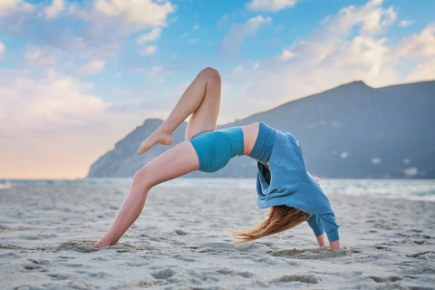 Woman practices Eka Pada Urdhva Dhanurasana on sandy beach while sun sets in the background,