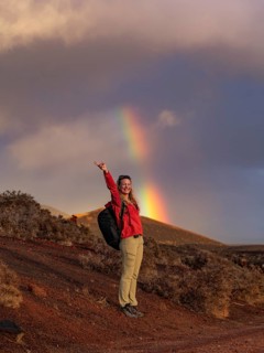 A hiker in a red jacket stands joyfully beneath a vibrant rainbow in Timanfaya National Park,