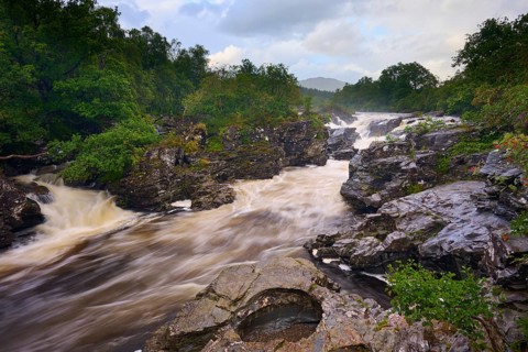 A wild river flows through a rocky landscape surrounded by lush greenery and cloudy skies, River
