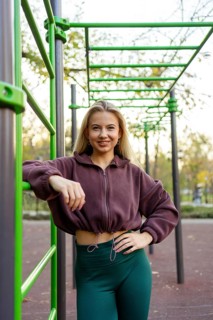 A confident woman in sportswear poses by green bars in an outdoor fitness park. She wears a maroon