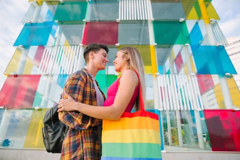 A happy lesbian couple shares a tender moment in front of a vibrant, colorful structure. They