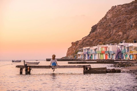 A lone woman sits on a pier in Klima, Milos Island, Greece, facing a serene sunset. Vibrant,