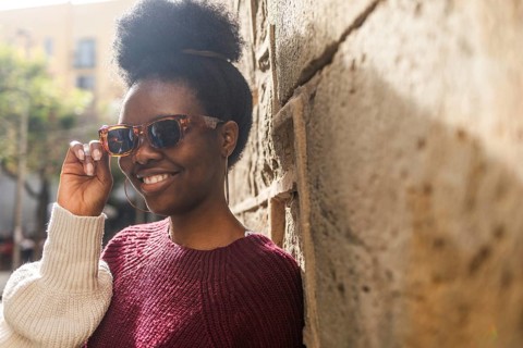 A fashionable Black woman wearing trendy sunglasses leans against a textured stone wall. Her