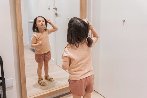 Young girl stands confidently before a mirror, brushing her hair with focus and intent. She's