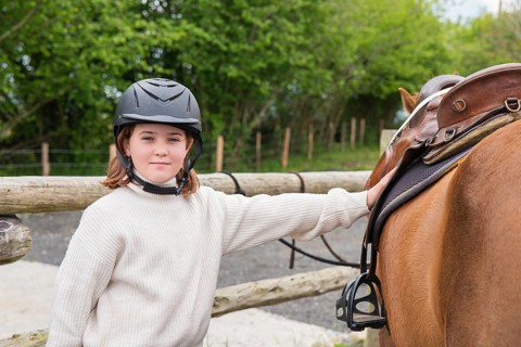 A young girl wearing a helmet stands beside her horse on a sunny farm day. The rural background of