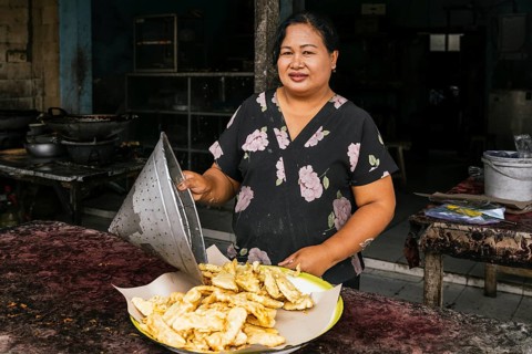 A street vendor stands behind a table displaying fresh fried snacks on a tray. She wears a floral