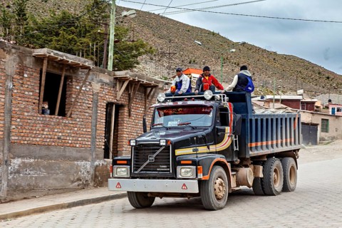 A truck drives through a village in a mountainous landscape, several people are sitting on the back