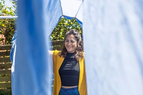 A woman stands outside, smiling as she hangs laundry on a clothesline. The sun shines brightly,