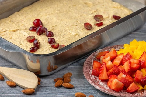 Unbaked dough in baking pan with grapes on top, strawberries and mango next to it