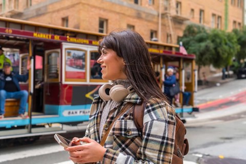 Smiling young woman exploring san francisco city, wearing headphones and backpack while holding a