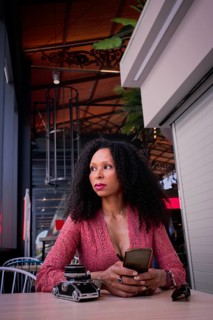 A latin woman sits at a cafe table, holding a smartphone. The ambient lighting creates a cozy