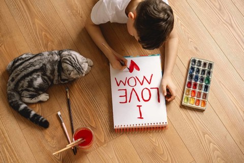 A boy lies on a wooden floor, painting a heartfelt Mother's Day card with I Love Mom. Nearby, a