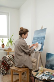 A focused artist paints on a canvas in her home studio, surrounded by natural light and cozy decor.