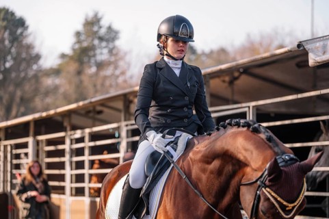An equestrian in full dressage attire, helmet, and gloves riding a bay horse at a stable The