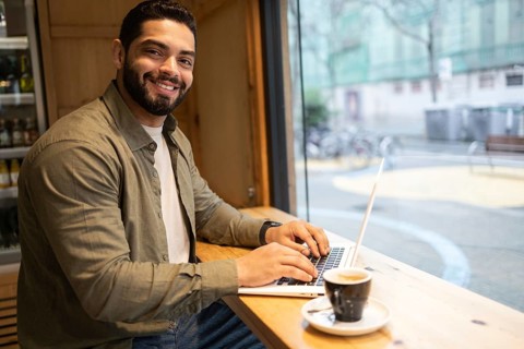 Man seated in a cozy cafe by the window, smiling and working on a laptop with a cup of coffee in
