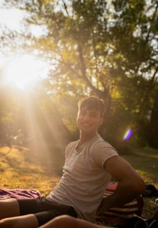 A cheerful young man enjoys the warm ambiance by the lake, sitting on a blanket with sunlight