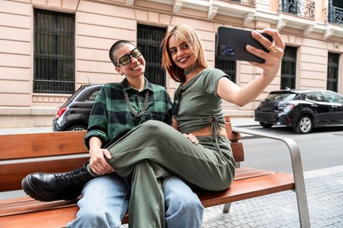 A joyful Colombian lesbian couple sitting on a bench, capturing a selfie. The image celebrates