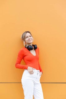 A woman in a red top and white pants leans against an orange wall in Warsaw, with headphones around