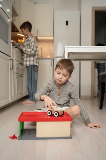 A young boy plays with a toy car on the kitchen floor, while his brother is engaged at the counter.
