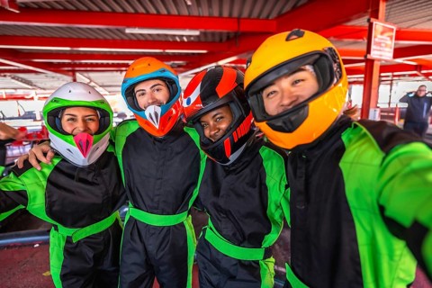Diverse friends wearing helmets and racing suits, smiling and taking a selfie together at an indoor