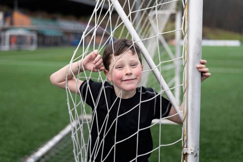 A joyful young boy playfully stands behind a soccer goal net on a lush green sports field. His