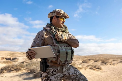A military professional stands in a desert environment, equipped with tactical gear, holding a
