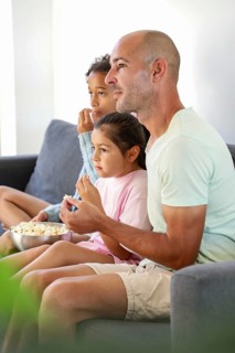 A family of three sits on a cozy sofa, engrossed in a movie while sharing a bowl of popcorn. The