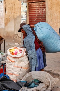 A woman wearing traditional clothes carries wool sacks over her shoulder in the street, The street