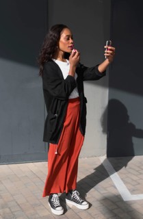 A young black woman in casual clothing applies lipstick outdoors using a compact mirror. She stands