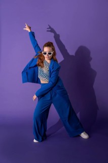 A young woman in a vibrant outfit dances energetically against a purple background. Her dynamic
