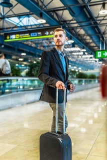 Businessman holding smartphone and pulling trolley suitcase while walking in airport terminal and