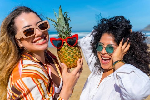 Two young women laughing on a sunny beach, holding a pineapple with heart sunglasses, enjoying a