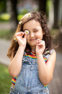 A cheerful, smiling little girl plays outdoors, holding a chamomile to her eye. Surrounded by
