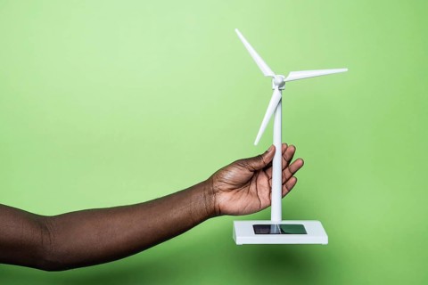 A hand holds a small wind turbine model against a green background, symbolizing sustainable energy