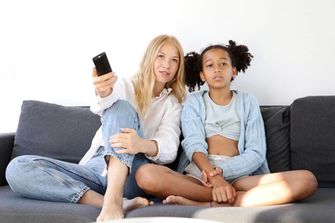 A young woman and a young girl sit on the couch holding a TV remote, watching a show together. The